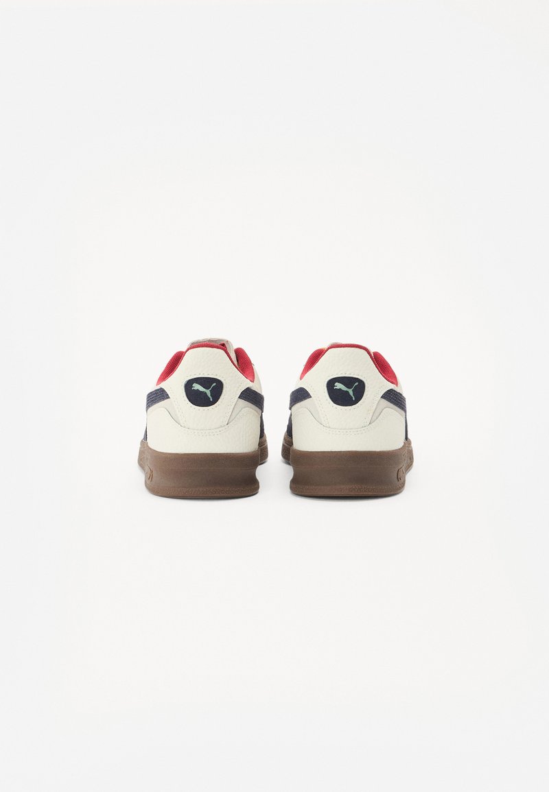 Białe sneakersy z czerwonymi i granatowymi akcentami, teksturowana skórzana cholewka oraz brązowa gumowa podeszwa. Logo PUMA widoczne z tyłu.
