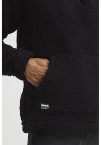 Pullover in pile nero con superficie texturizzata, tasca frontale ampia e polsini a coste. Presenta un piccolo tag con il marchio vicino all'orlo.