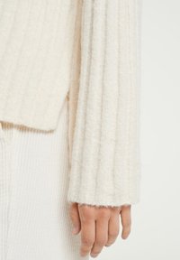 Pull en tricot ivoire avec texture côtelée verticale ; tissu doux et duveteux ; manche large se terminant par un ourlet roulé ; main posée contre le tissu.