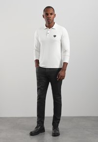 Weißes langärmeliges Poloshirt mit Frontlogo, kombiniert mit schwarzen Hosen und schwarzen Schnürstiefeln. Schlichtes Design, glatte Textur und taillierte Passform.