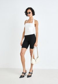 Haut à bretelles blanc avec fronces, short de cyclisme noir, escarpins noirs à talons aiguilles avec accents de boucle, et sac à épaule blanc. Textures lisses, design ajusté.