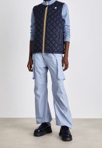 Marineblaue wattierte Weste mit gelbem Reißverschluss, kombiniert mit hellblauen Cargo-Hosen mit Seitentaschen und weitem Schnitt. Schwarze Plateau-Schuhe.