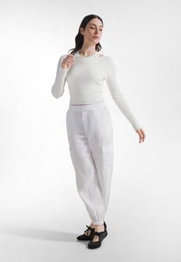 Weißes, geripptes Crop-Top mit langen Ärmeln und freiliegenden Schultern, kombiniert mit hellcremefarbenen Cargo-Hosen mit elastischen Bündchen und Seitentaschen. Schuhe mit schwarzer Schleife als Akzent.