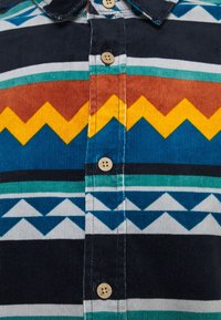Camisa abotonada con una tela de corduroy estampada en zigzag de colores azul, amarillo, marrón y verde, con diseños geométricos.