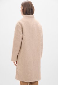 Beige Mantel aus strukturiertem, plüschigem Stoff, mit hohem Kragen und langen Ärmeln, von hinten vor einem hellen Hintergrund gezeigt.