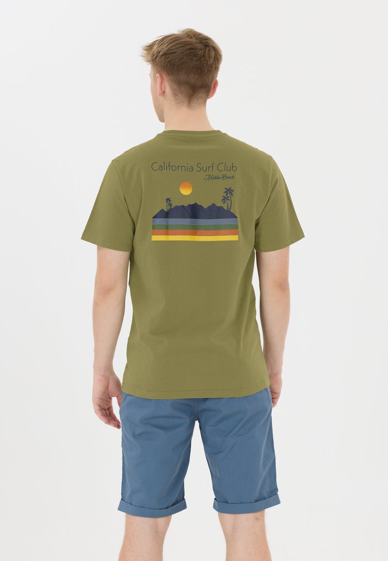 Olivi vihreä T-paita, jossa on graafinen printti vuorista, palmuista ja auringosta, sekä vaakasuorat raidat keltaisena, sinisenä ja vihreänä.