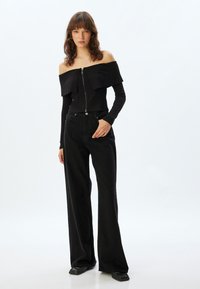Schwarzes, geripptes Off-Shoulder-Oberteil mit Reißverschluss, begleitet von weit geschnittenen, hoch taillierten schwarzen Jeans und strukturierten Schuhen.