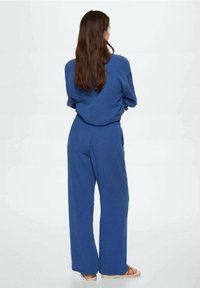 Sininen puuvillainen asu, jossa on löysä pitkähihaisen yläosan ja leveälahkeiset housut. Kankaassa on teksturoidun pinnan viimeistely. Jalat paljasti sandaalit.