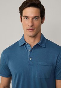 Pikétröja i mörkblått, tillverkad av mjuk bomull. Har en knappslå med två knappar, krage och en framficka med diskreta sömdekor.