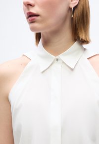 Γυναίκα με κοντά ανοιχτό καστανά μαλλιά φοράει λευκό αμάνικο πουκάμισο με γιακά και χρυσό κρίκο στο αυτί, κοιτάζοντας ελαφρώς προς το πλάι.