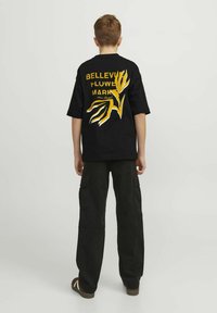 Juodas marškinėliai su geltonu grafiniu dizainu, vaizduojančiu rankas ir tekstą "BELLEVUE FLOWER MARK" ant nugaros, derinami su juodomis kargo kelnėmis.