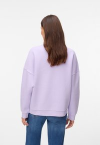 Femme aux longs cheveux bruns portant un sweat-shirt violet clair et un jean bleu, debout de dos face à la caméra.