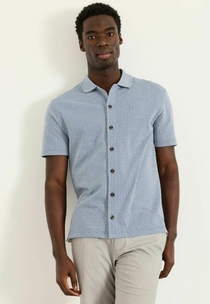 Mann trägt ein hellblaues, kurzärmeliges Hemd mit kleinem geometrischem Muster und hellgraue Hose, steht an einer weißen Wand.