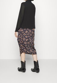 Donna che indossa un top lungo a maniche trasparenti nero, una gonna midi leopardata e stivali da cowboy neri, in piedi contro uno sfondo bianco semplice.