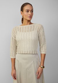Hellbeige gestrickter Pullover mit lockerem, offenem Maschenbild, Dreiviertelärmel und weitem Ausschnitt, kombiniert mit einem hellen Rock.