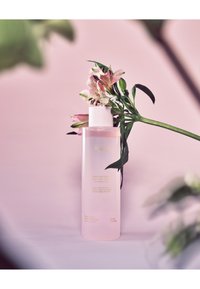 Huxley toner i en klar, cylindrisk flaska, rosa vätska, vit kork. Texten visar produktnamn och detaljer. Blomsterdesign i bakgrunden.