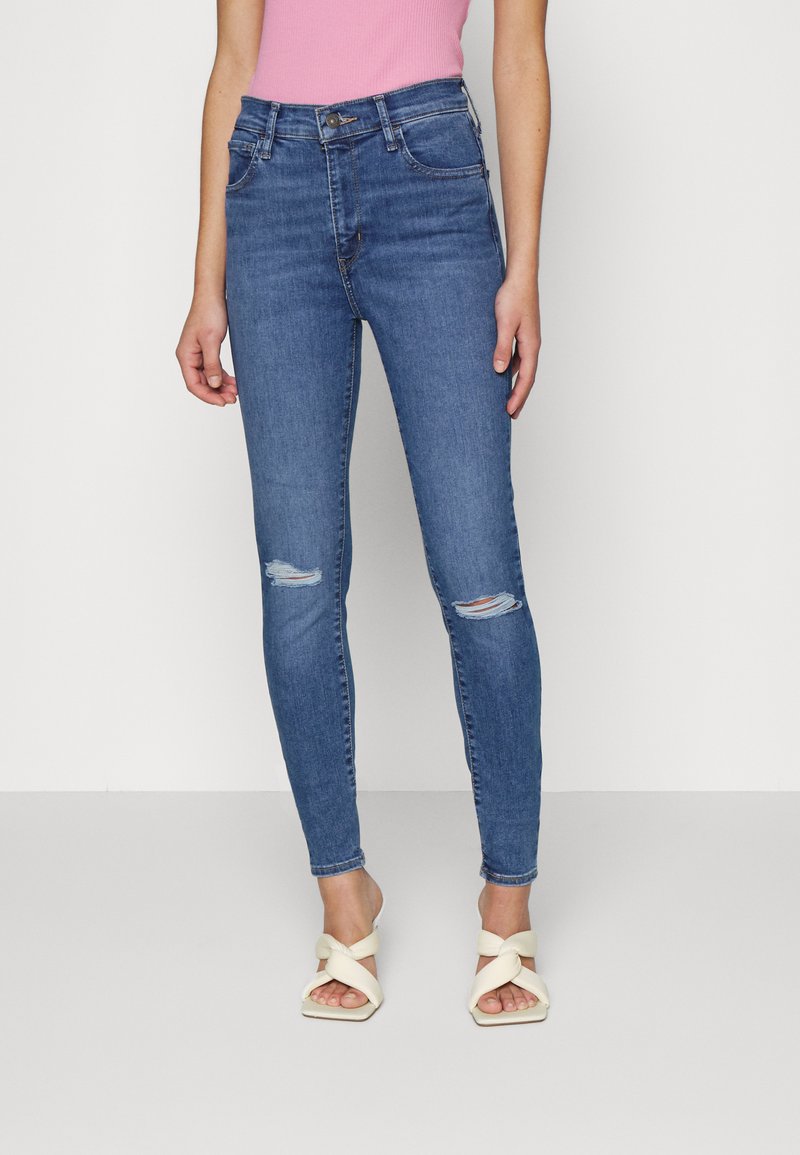 Jeans de denim azul ajustados con detalles desgastados en las rodillas, diseño de tiro alto y textura suave, combinados con sandalias blancas de tiras.