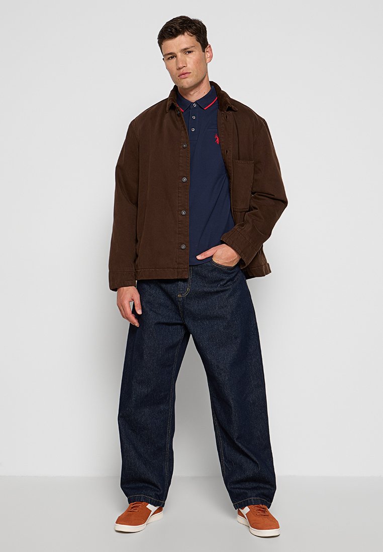 Braune Kordjacke über einem marineblauen Polo mit roten Akzenten, kombiniert mit lockeren blauen Jeans und orangefarbigen Sneakers. Frontalansicht, neutraler Hintergrund.
