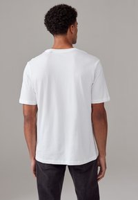 Fehér pamutpóló laza fazonnal, kerek nyakkal, rövid ujjakkal és puha textúrával, hátulról egy semleges háttér előtt bemutatva.