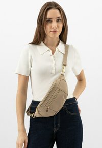Marsupio in pelle beige con molteplici scomparti con zip, tracolla regolabile, texture liscia e dettagli in oro, indossato sopra jeans scuri.