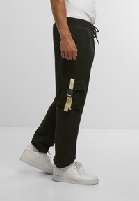 Mann med svarte cargo-bukser med beige stroppdetaljer og hvite sneakers, som står mot en enkel lys grå bakgrunn.