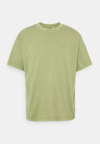 T-shirt en coton vert avec un col rond, des manches courtes et des fentes sur les côtés. Présente un logo subtil brodé sur la poitrine. Texture douce.
