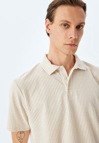 Polo shirt beige chiaro a maniche corte con righe verticali, colletto classico e chiusura a tre bottoni. Tessuto morbido con vestibilità rilassata.