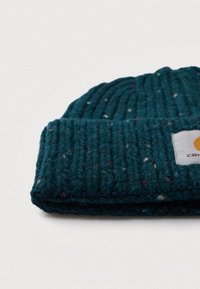 Berretto lavorato a maglia di un verde scuro con risvolto e piccolo patch rettangolare bianco su sfondo chiaro.