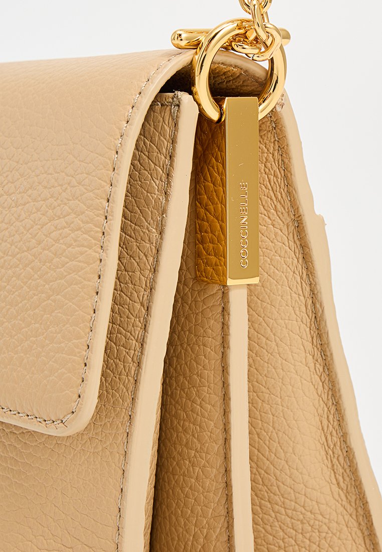 Borsa a tracolla in pelle beige con finitura testurizzata. Presenta hardware dorato, un dettaglio a catena e un'etichetta rettangolare incisa con "COCCINELLE."