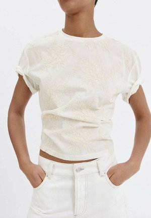 Personne portant un haut en dentelle crème à manches courtes à motifs floraux, poignets roulés, et un jean blanc taille haute, les mains dans les poches devant un fond uni.