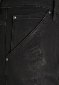 Černé džínsy z denimu s boční kapsou, kovovými nýty, strukturovanými švy a texturovaným povrchem.