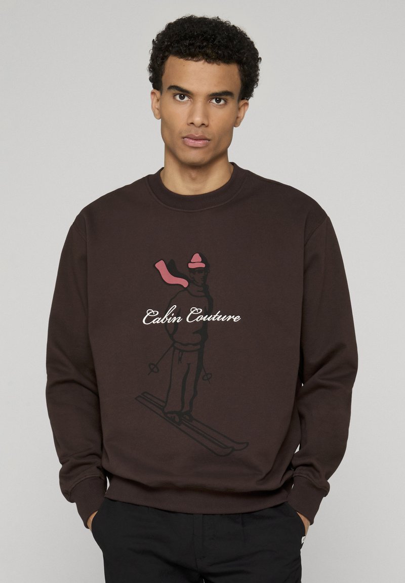 Sweat-shirt en coton marron avec un motif de skieur en noir et rose, texte "Cabin Couture" en dessous, col et poignets côtelés.
