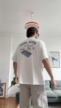 Biela bavlnená tričko s textom "POOL CLUB" a grafikou bazénovej scény na zadnej strane; voľný strih a krátke rukávy, skombinované s svetlosivými šortkami.
