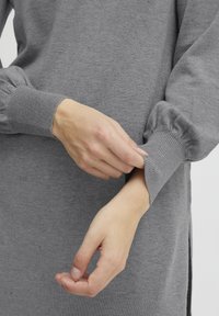 Grauer Strickpullover mit gerippten Bündchen und lockerer Passform. Verfügt über einen seitlichen Schlitz und eine weiche Textur. Hand, die das Bündchen anpasst.