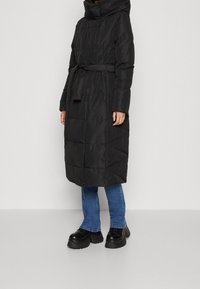 Kvinna klädd i en lång svart vadderad kappa med huva och bälte, blå jeans och svarta platåskor mot en enkel ljus bakgrund.