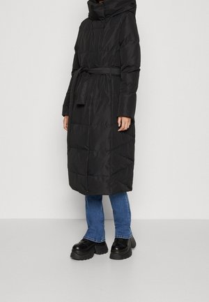 Frau in einem langen schwarzen gesteppte Mantel mit Kapuze und Gürtel, blauen Jeans und schwarzen Plateauschuhen vor einem schlichten hellen Hintergrund.