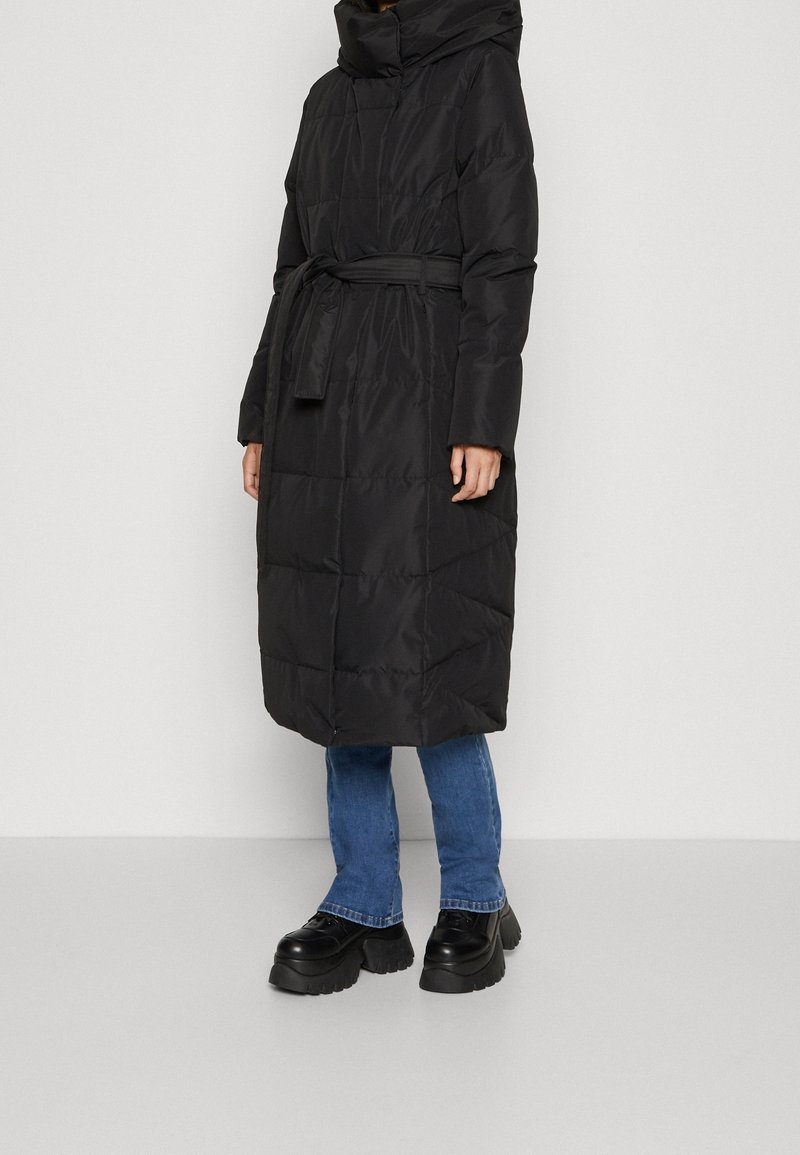 Kvinna klädd i en lång svart vadderad kappa med huva och bälte, blå jeans och svarta platåskor mot en enkel ljus bakgrund.