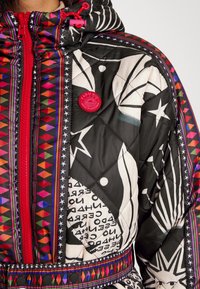 Gequilte jas met een zwart-wit geometrisch ontwerp en kleurrijke patronen, een felrode rits en een opvallende logo patch.