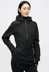 Jovem mulher vestindo uma jaqueta preta ajustada com capuz, cinto na cintura e bolsos com zíper, de pé com a mão no bolso contra um fundo simples.