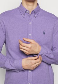 Fialová košile s dlouhým rukávem, zapínáním na knoflíky a klasickým límcem, s jemnou texturou a vyšitým logem na levé hrudi.