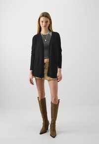 Svart strikket cardigan med åpen front og lommer, brukt over en grå kort topp og et beige miniskjørt; kombinert med brune knelengde støvler.