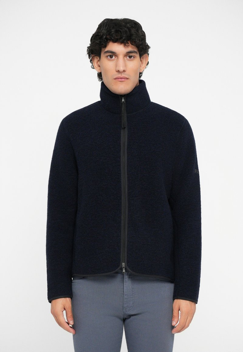 Marineblaue Fleecejacke mit hohem Kragen, durchgehendem Reißverschluss, schwarzem Kontrastreißverschluss und subtiler Textur. Getragen über hellgrauen Hosen.