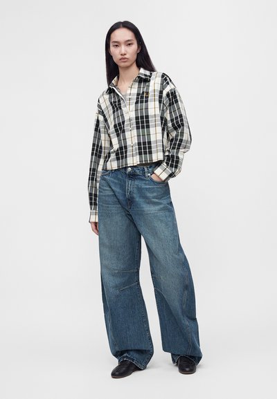 Mujer con cabello largo y oscuro, que lleva una camisa de cuadros blanca y negra y vaqueros azules holgados, de pie con una mano en el bolsillo.