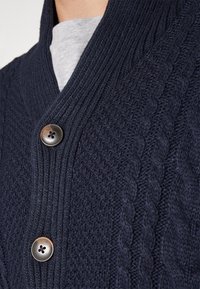 Námořnicky modrý pletený kardigan s texturovaným copánkovým vzorem, šálovým límcem a dvěma hnědými knoflíky vpředu. Pod ním je vidět šedé tričko.
