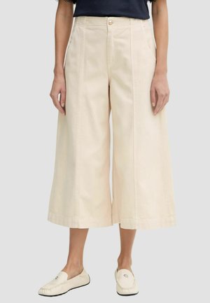 Donna che indossa pantaloni crema a vita alta e gamba larga, corti con dettaglio bottoni, top nero e mocassini crema su sfondo neutro.