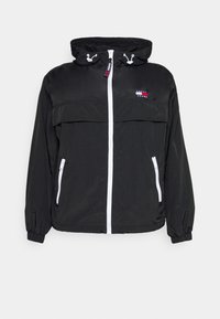 Svart vindjacka med huva, med en vit dragkedja fram, två sidofickor med dragkedja och en logotyp på bröstet. Slät textur.