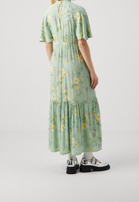 Blumiges Maxikleid in Hellgrün mit gelben und lila Blumen. Verfügt über kurze Rüschenärmel und einen gestuften Rock. Kombiniert mit silbernen Plateauschuhen.