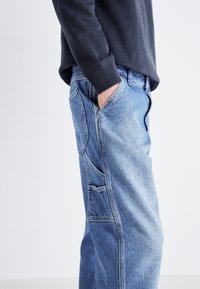 Light blue denim jeans with a relaxed fit, featuring a side pocket, textured fabric, and a mid-rise waist. Paired with a dark long-sleeve shirt.
