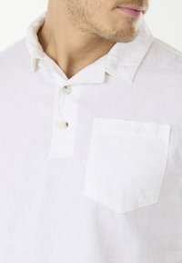 Weißes Leinenhemd mit einem geknöpften Kragen, drei Knöpfen am Hals und einer kleinen Brusttasche mit dezentem Branding.