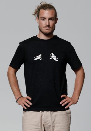 Schwarzes Baumwoll-T-Shirt mit zwei weißen Astronauten-Grafiken und bunten Akzenten, vor einem gefleckten, sternenklaren Hintergrund.